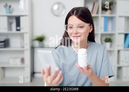 Jeune femme médecin en uniforme et lunettes tenant smartphone et montrer la bouteille en plastique contenant un nouveau médicament efficace au patient en ligne Banque D'Images