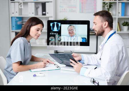 Deux praticiens assis devant l'écran d'ordinateur et discutant une image radiologique tout en la montrant au médecin senior pendant la connexion communication Banque D'Images