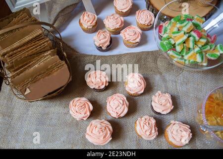 Les petits gâteaux roses à la vanille et au chocolat dépoli sont placés sur une nappe de toile de jute avec d'autres bonbons à l'arrière-plan comme table de dessert pour un mariage. Banque D'Images