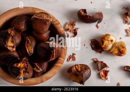 Un bol en bois de châtaignes grillées maison au four avec des coquilles et des peaux éparpillées sur le plateau en marbre. Une collation traditionnelle d'hiver, d'automne avec Banque D'Images