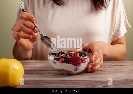 Une femme caucasienne mange un bol en verre frais fait maison de yaourt crémeux parfait avec des baies, muesli et des graines dans elle sur table en bois gros plan Banque D'Images