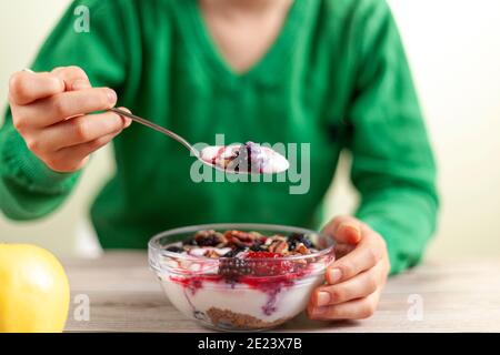 Un enfant avec une chemise verte mange un bol en verre frais fait maison de yaourt crémeux parfait avec des baies, du muesli et des graines sur une table en bois gros plan photo Banque D'Images