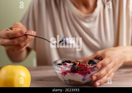 Une femme caucasienne mange un bol en verre frais fait maison de yaourt crémeux parfait avec des baies, muesli et des graines dans elle sur table en bois gros plan Banque D'Images