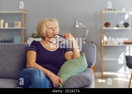 Femme mûre détendue assise sur un canapé, tenant des lunettes, regardant loin et pensant Banque D'Images