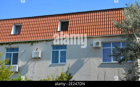 Une petite maison résidentielle avec un toit en argile rouge mansarde et des fenêtres mansarde ou mansarde, air conditionné unités extérieures sur un mur gris stuc. Banque D'Images