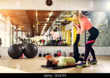 Belle jeune femme instructeur aidant sa cliente à étirer ses jambes après un entraînement difficile dans une salle de gym. Banque D'Images