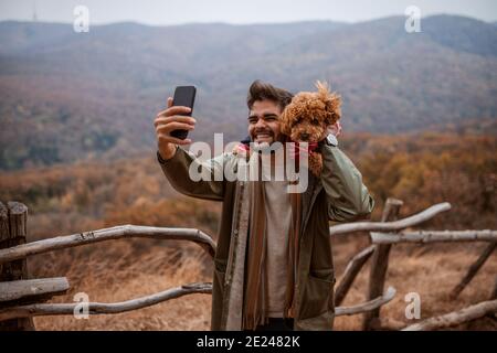 Homme prenant autoportrait avec le chien sur les épaules dans la nature. En arrière-plan montagnes et forêt. Heure d'automne. Banque D'Images
