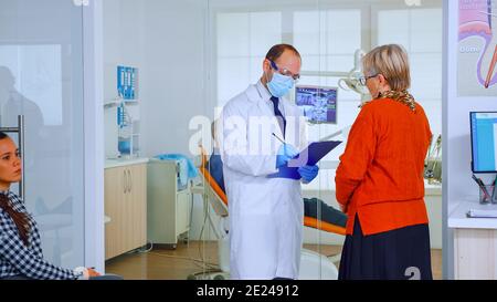 Orthodontiste avec masque parlant avec une femme âgée debout dans la salle d'attente de la clinique stomatologique prenant des notes sur le presse-papiers. Infirmière tapant sur des rendez-vous informatiques dans un bureau moderne bondé Banque D'Images