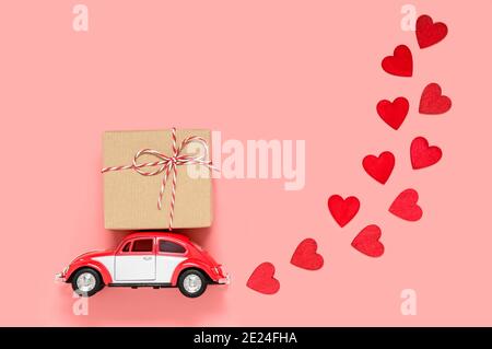 Fulda, Allemagne - DEC 18, 2020: Petite voiture de jouet rétro rouge livrant boîte cadeau pour la Saint-Valentin sur fond rose avec de nombreux coeurs rouges. Amour et holidés Banque D'Images