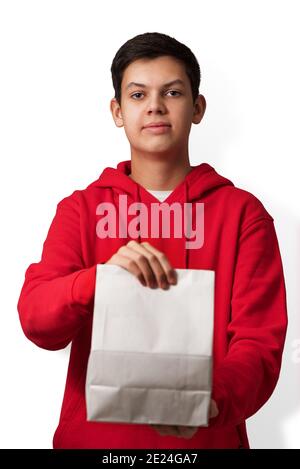 Adolescent tenant un sac de papier blanc dans sa main isolé sur fond blanc. Jeune homme caucasien portant le sweat à capuche rouge offrant le paquet avec le contenu Banque D'Images