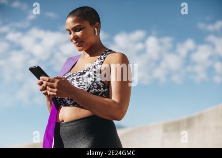 Femme en vêtements de sport utilisant son smartphone après une séance d'entraînement. Une femme en bonne santé, portant des écouteurs sans fil, prend une pause après son entraînement et le vérifie Banque D'Images