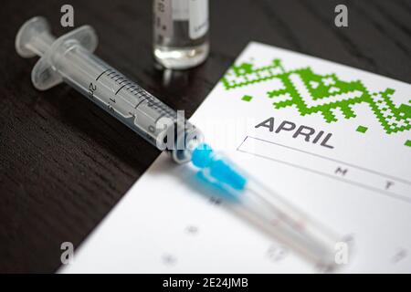 Gros plan d'une seringue médicale placée sur un calendrier d'avril avec un flacon de vaccin en arrière-plan, symbolisant la vaccination programmée Banque D'Images