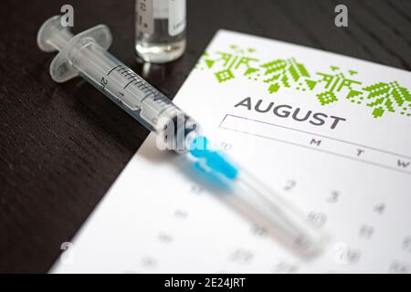 Gros plan d'une seringue médicale placée sur un calendrier d'août avec un flacon de vaccin en arrière-plan, symbolisant la vaccination programmée Banque D'Images