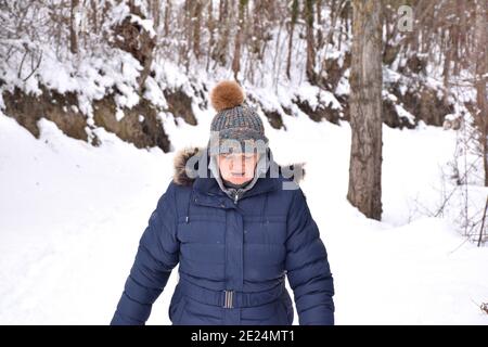 Femme à la retraite en veste et chapeau avec des couleurs vives regardant où elle marche sur un chemin rempli de neige dans la forêt. Scène après la tempête de neige appelée Filom Banque D'Images