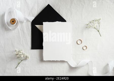 Maquette de carte d'invitation de mariage. Carte vierge en papier blanc, enveloppe noire, anneaux dorés, ruban, fleurs vue du dessus. Banque D'Images