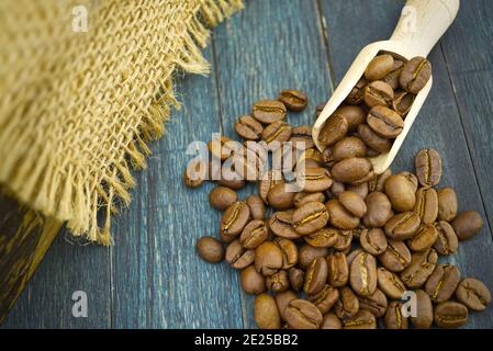 Grains de café torréfiés qui se répande d'une cuillère en bois sur du bois et un arrière-plan en tissu hessien ou en toile de jute Banque D'Images