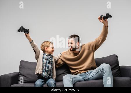 KIEV, UKRAINE - 17 NOVEMBRE 2020 : père et fils joyeux montrant un geste de victoire avec des joysticks isolés sur gris Banque D'Images