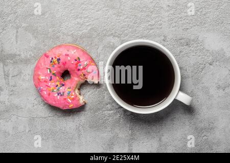 Donut rose glacé avec saupoudrer et une morsure prise dehors à côté d'une tasse de café noir avec vue sur la pose à plat au plafond Banque D'Images