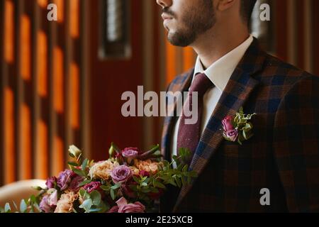 Portrait court d'un beau homme méconnaissable, dans une cravate et un élégant costume à carreaux avec une boutonnière, tenant un grand bouquet de fleurs fraîches Banque D'Images