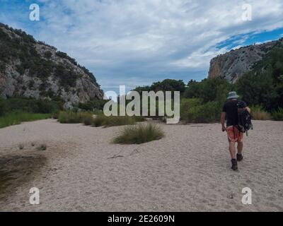 Homme touriste marchant sur le chemin du sable à la plage de Cala Luna au golfe d'Orosei avec des buissons verts, des arbres et des roches calcaires. Sardaigne, Italie. Banque D'Images