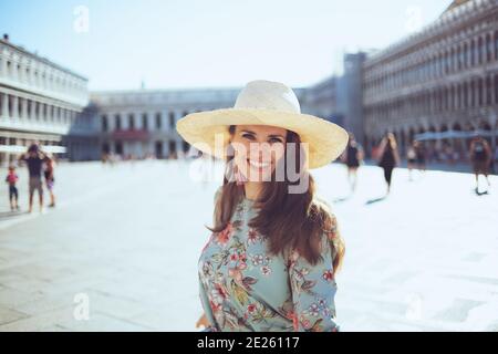 Jeune voyageur solo souriant femme en robe florale avec chapeau ayant une visite à pied à la place San Marco à Venise, Italie. Banque D'Images