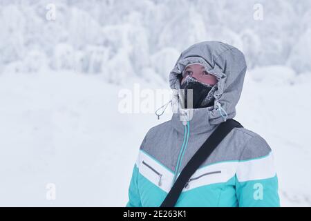 jeune femme dans une veste chaude avec une capuche et un soufflet de cou de chamboule sur son visage contre l'arrière-plan de paysage d'hiver flou Banque D'Images