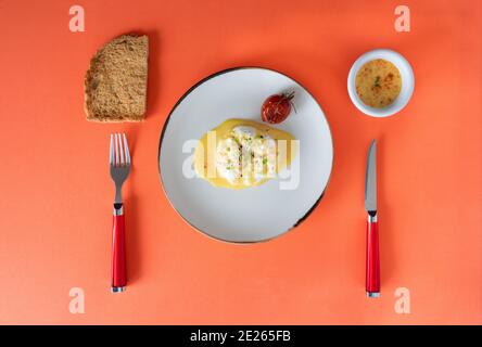 table de petit-déjeuner oeuf poché avec sauce dans un petit bol ciboulette hachée pain grillé plaque blanche tomate rôtie sur fond orange. épices, poivre noir Banque D'Images