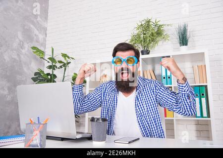 Un homme à barbe heureux célèbre la victoire en faisant un geste gagnant sur un bureau dans un bureau moderne, célébration. Banque D'Images