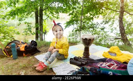 Une charmante fille sourit et s'assoit sur une couverture dans un camping familial, entouré d'instruments de musique, de plats, de sacs à dos. Reposez-vous dans la forêt pendant un tr Banque D'Images