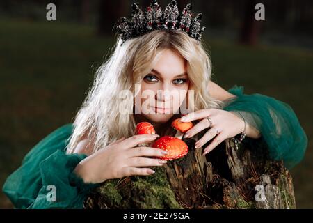 Une belle jeune femme blonde dans une longue robe verte et un diadem sur sa tête dans la forêt. Fille assise près de la vieille souche avec amanitas. Glar solaire Banque D'Images