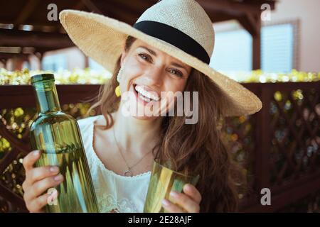 Portrait de la femme au foyer élégante et heureuse en chemise blanche avec bouteille d'eau, verre et chapeau sur la terrasse. Banque D'Images