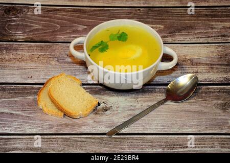 Une assiette profonde de bouillon de poulet chaud avec un œuf dur et une branche de persil sur une table en bois, à côté d'une cuillère et de deux tranches de pain de blé. Côté vie Banque D'Images