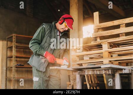 Image de l'homme mûr charpentier dans le bois de façonnage uniforme de protection avec broyeur électrique. En appréciant son travail dans son garage de travail le jour ensoleillé. Banque D'Images