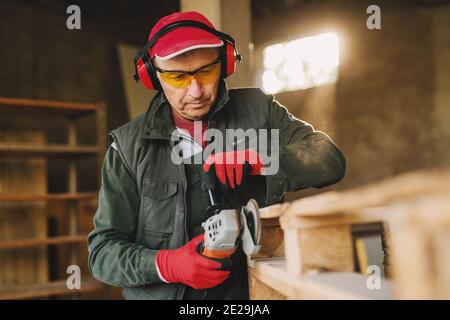 Image de l'homme mûr charpentier dans le bois de façonnage uniforme de protection avec broyeur électrique. En appréciant son travail dans son garage de travail le jour ensoleillé. Banque D'Images
