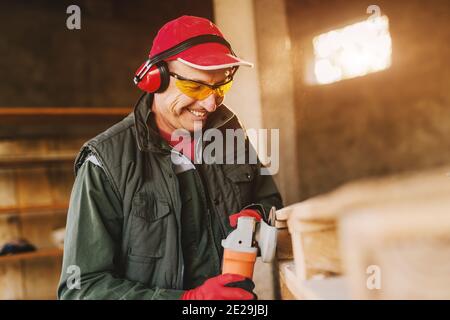 Photo de l'homme mûr et souriant de menuisier en uniforme de protection façonnant le bois avec moulin électrique. En appréciant son travail dans son garage de travail le jour ensoleillé. Banque D'Images