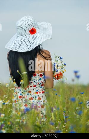 Fille dans une robe et un chapeau blanc se tient dans un champ avec un bouquet de fleurs sauvages Banque D'Images