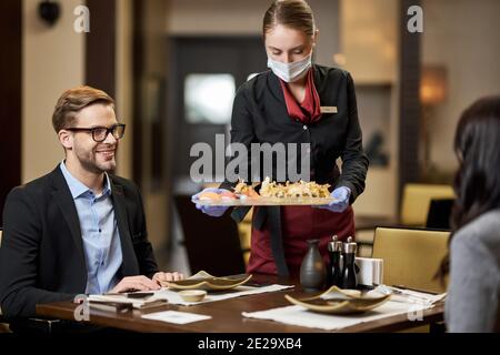 Un jeune homme content dînant avec une dame pendant qu'il était professionnel serveuse sous masque servant des sushis Banque D'Images