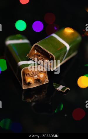 Barre de chocolat au lait avec arachides, biscuits et caramel salé sur fond de miroir noir. Des lumières de Noël dans le reflet Banque D'Images