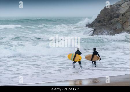 Surfeurs transportant leurs planches de surf et courant dans une mer glaciale pendant les conditions météorologiques hivernales froides sur la plage Great Western à Newquay, en Cornouailles. Banque D'Images