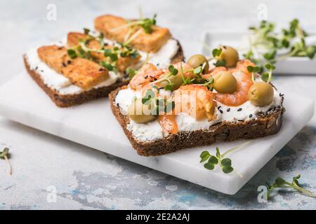 Variété de sandwiches pour le petit déjeuner, en-cas, hors-d'œuvre - tempeh, saumon, crevettes, sandwichs au pain aux grains entiers grillés avec des micro-légumes sur un fond léger Banque D'Images