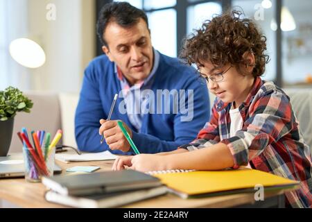 Un père latin d'âge moyen, soucieux de s'occuper de ses devoirs avec son fils et son enfant, tout en étant assis à son bureau à la maison. Étude en ligne, famille, concept de paternité Banque D'Images