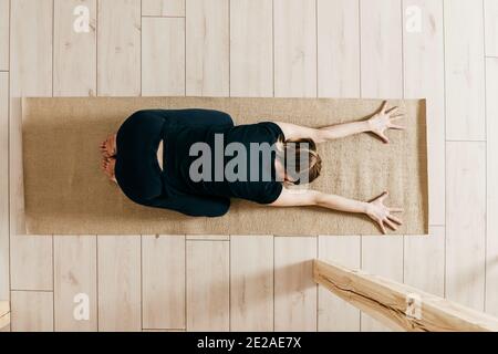 Vue de dessus de femme pratiquant le yoga à l'intérieur. La femme se courbe et s'étire vers l'avant. Femme de fitness sur tapis d'exercice sur des sols clairs. Banque D'Images