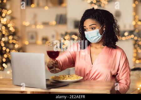Femme afro en masque de boire du vin pendant la vidéo sur ordinateur portable Banque D'Images