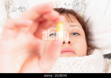 une jeune fille enveloppée d'un foulard est malade à la maison, se trouve dans le lit et tient une capsule avec des médicaments dans sa main Banque D'Images
