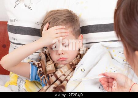 un petit garçon un enfant enveloppé dans un foulard est malade à la maison, repose dans le lit et tient sur son front, maman lui donne un thermomètre Banque D'Images