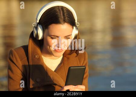 Femme satisfaite écoutant de la musique avec un casque et un téléphone portable sur la plage au coucher du soleil en hiver
