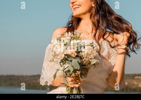 belle fille mince avec de longs cheveux bouclés dans une robe en dentelle. femme tenant un bouquet de fleurs et riant contre le ciel Banque D'Images