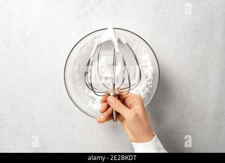 Main de jeune femme tenant le fouet sur le bol de crème fouettée. Fond blanc de cuisine. Cuisine et concept culinaire maison. Style de pose à plat. Banque D'Images