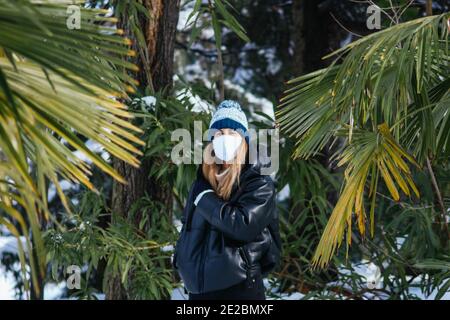 Photo d'une jeune femme dans un visage avec mise au point peu profonde masque et chapeau tricoté entouré de palmiers Banque D'Images