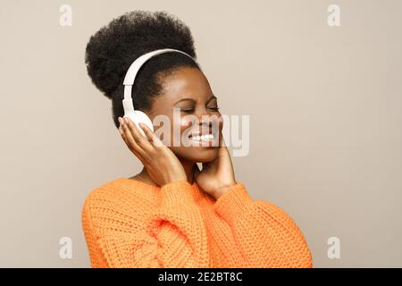 Femme afro souriante dans un cavalier orange touchant un casque sans fil, écoute de la musique préférée isolée Banque D'Images